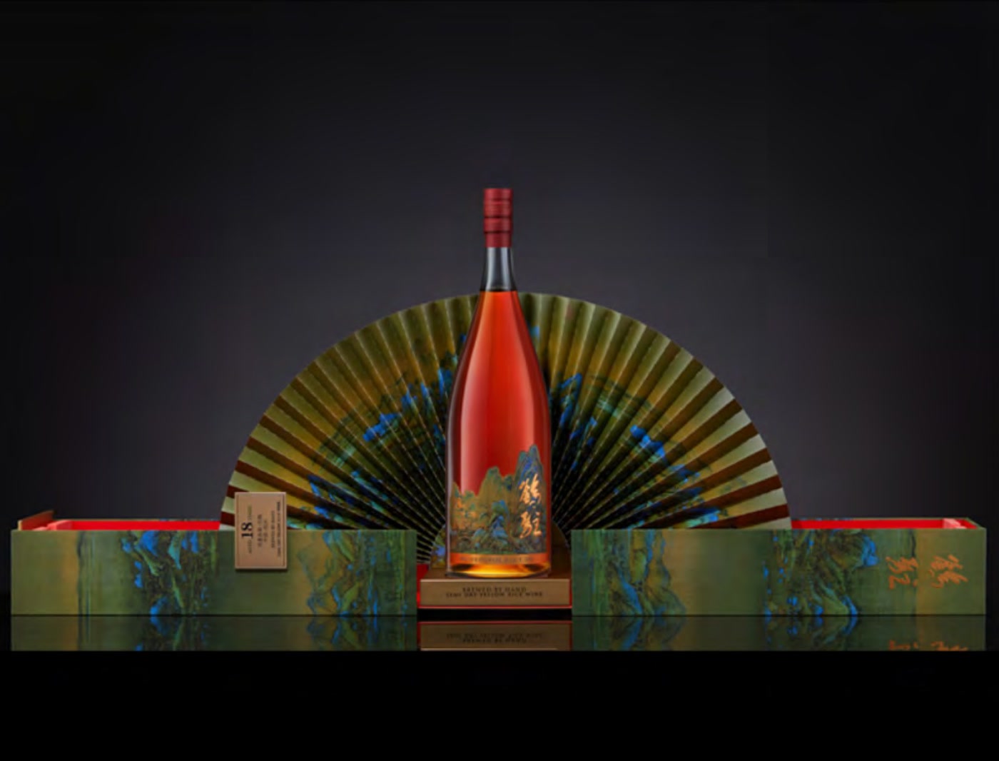 A bottle standing in front of an opened blue-green landscape fan, flanked by open gift boxes with red lining, against a dark background