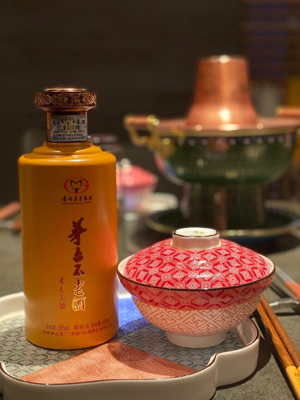 皿の横に立つ瓶。右手に蓋つきの磁器の碗、背景は暖色の照明の宴席